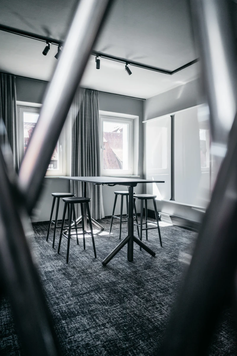 Moderner Besprechungsraum mit hohen Bartischen und Hockern auf grauem Teppichboden. Gro&szlig;e Fenster mit Vorh&auml;ngen und Deckenspots sorgen f&uuml;r helle, elegante Atmosph&auml;re. Blick durch einen verschwommenen Vordergrund, der die Tiefe des Raumes betont.