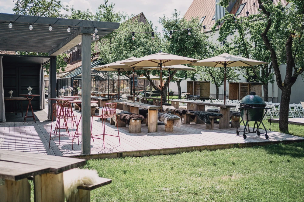 Sonnige Terrasse mit Holztischen, wei&szlig;en Sonnenschirmen und Sitzgelegenheiten. Gr&uuml;ner Rasenbereich und ein &uuml;berdachter Bereich mit roten Barhocker-St&uuml;hlen, umgeben von B&auml;umen und Geb&auml;uden.