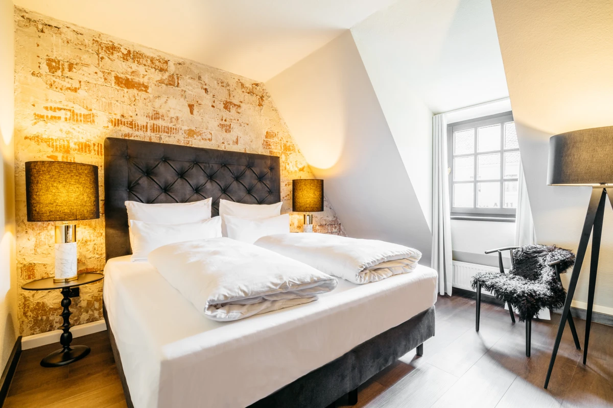 Gem&uuml;tliches Hotelzimmer mit gepolstertem Kopfteil und wei&szlig;er Bettw&auml;sche, einem charaktervollen Akzentstein an der Wand und warmer Beleuchtung durch seitliche Tischlampen. Gro&szlig;es Fenster l&auml;sst nat&uuml;rliches Licht herein, ein gepolsterter Sessel mit Fellauflage vervollst&auml;ndigt die einladende Atmosph&auml;re.