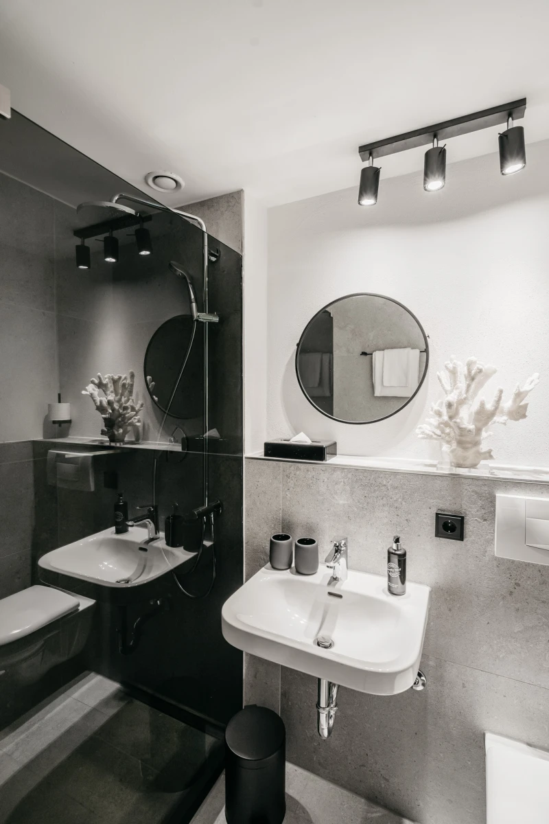 Modernes Badezimmer in Grau- und Wei&szlig;t&ouml;nen mit zwei Waschbecken, runden Spiegeln und minimalistischer Beleuchtung. Schwarze Armaturen und Accessoires kontrastieren mit den hellen W&auml;nden und verleihen dem Raum einen eleganten, zeitgen&ouml;ssischen Charakter.