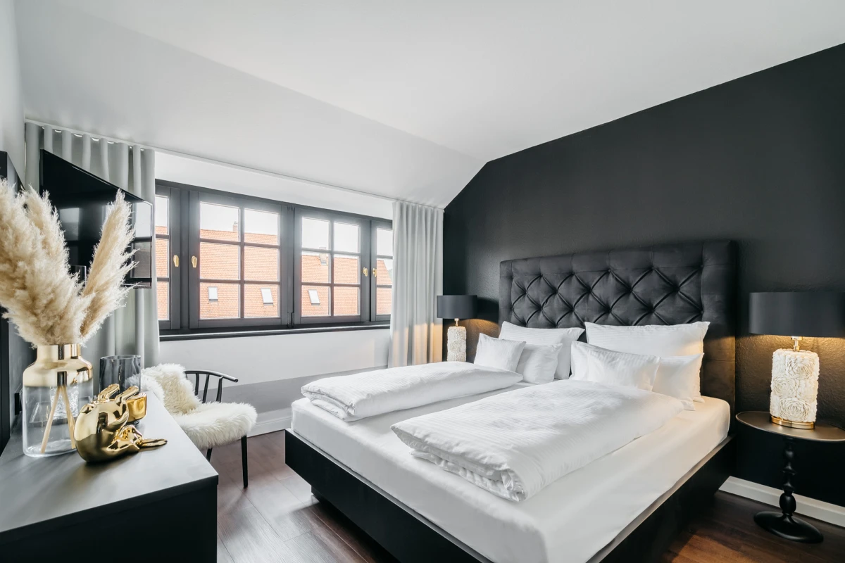 Modernes Hotelzimmer mit gro&szlig;er Fensterfront und anthrazitfarbener Akzandwand. Komfortables Bett mit gestepptem Kopfteil, dekoriert mit wei&szlig;er Bettw&auml;sche und Zierkissen. Stilvolle Dekoration mit Pampasgras in Glasvase und goldfarbenen Accessoires auf dem Nachttisch.