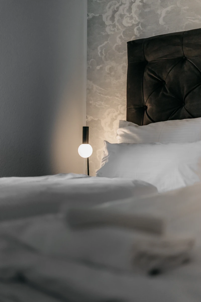 Minimalistisches Hotelzimmer mit gepolstertem Kopfteil in dunkler Farbe und wei&szlig;er Bettw&auml;sche. Eine moderne Stehlampe mit kugelf&ouml;rmigem Licht steht neben dem Bett, das dezent beleuchtet wird. Die Atmosph&auml;re ist ruhig und elegant mit ged&auml;mpften Graut&ouml;nen.