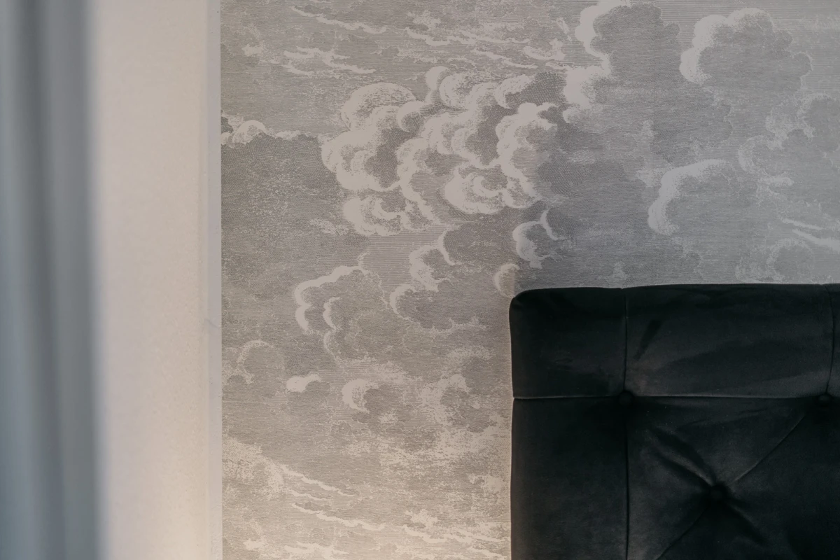 Wolkenf&ouml;rmige Tapete in ged&auml;mpften Graut&ouml;nen mit weichen, geschwungenen Wolkenformen. Im Vordergrund ein Ausschnitt eines schwarzen gepolsterten Sitzm&ouml;bels, vermutlich Teil eines Sessels oder Sofas.