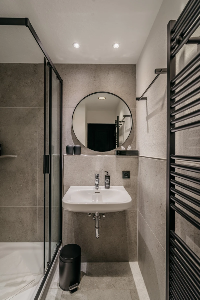 Modernes, minimalistisches Badezimmer mit grauen Wandfliesen, rundem Spiegel und wei&szlig;em Waschbecken. Eine Glasdusche und ein schwarzer Papierkorb erg&auml;nzen die schlichte Einrichtung, w&auml;hrend ein schwarzer Handtuchhalter an der Wand montiert ist.