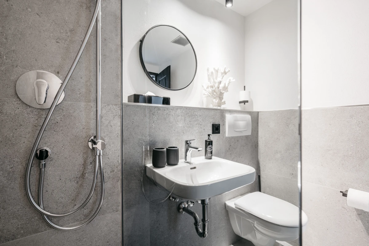 Modernes, minimalistisches Badezimmer mit grauen W&auml;nden, wei&szlig;er Toilette, rundem Spiegel und Dusche mit Brausekopf. Waschbecken mit schwarzen Accessoires auf einem grauen Regal montiert, helle, schlichte Atmosph&auml;re.