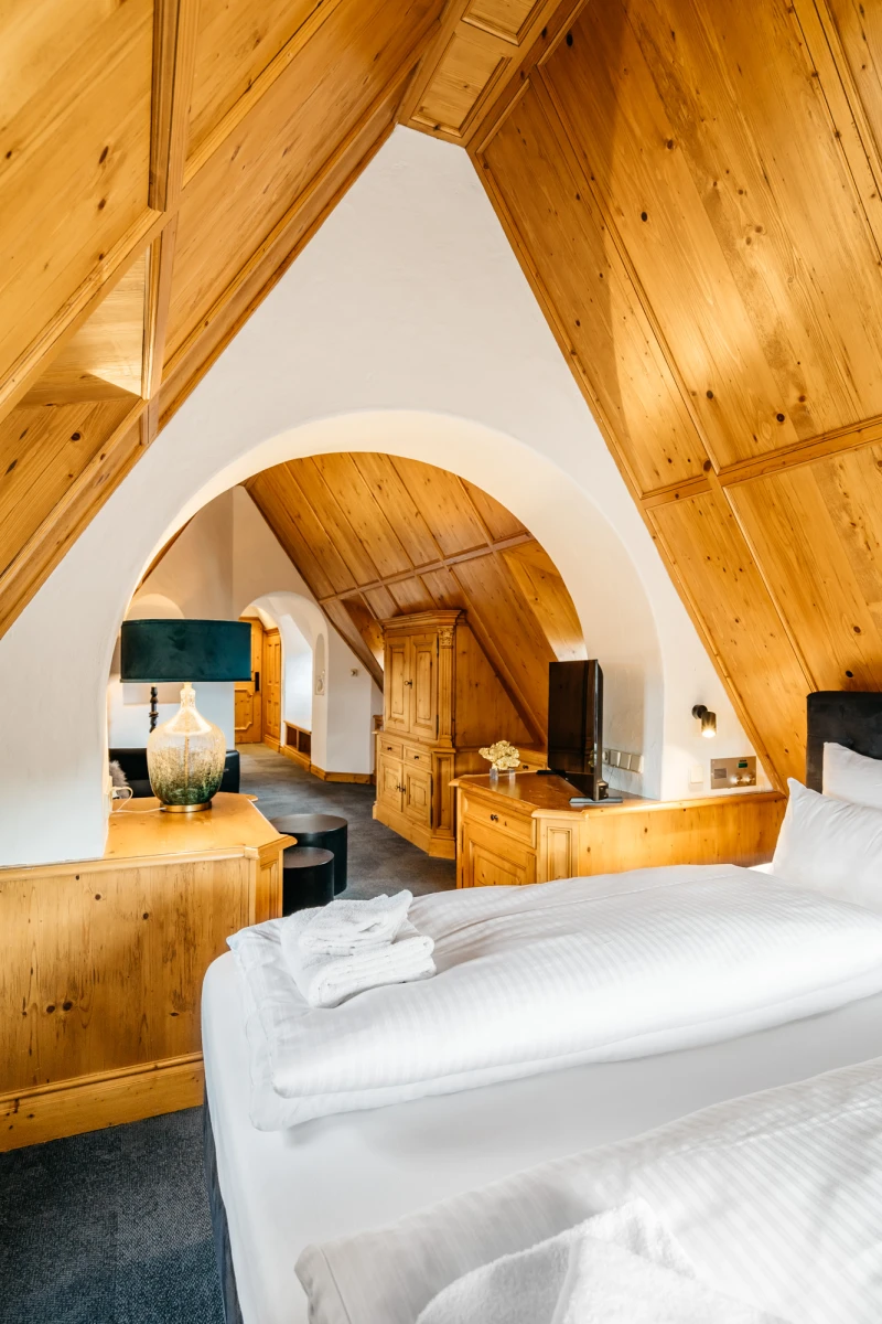 Gem&uuml;tliches Dachzimmer mit Holzvert&auml;felungen, wei&szlig; bezogenen Betten und einem geschwungenen wei&szlig;en Bogen, der verschiedene Bereiche des Raumes verbindet. Rustikale Holzm&ouml;bel und warme Holzt&ouml;ne schaffen eine einladende Atmosph&auml;re unter der schr&auml;gen Dachdecke.