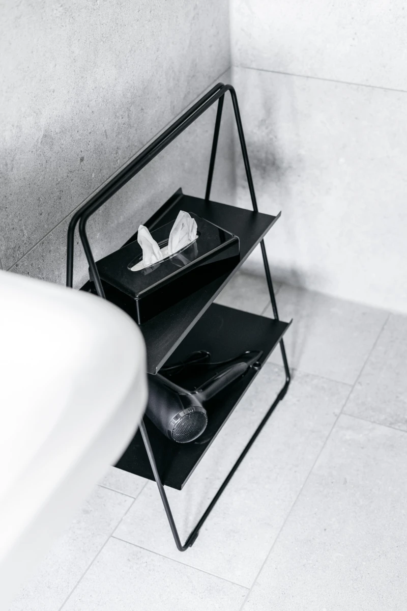 Modernes, minimalistisches schwarzes Regal mit zwei Ebenen, auf dem eine wei&szlig;e Tissuebox und ein schwarzer Ball liegen. Das Regal steht in einer monochromen, hellen Ecke eines Raumes.